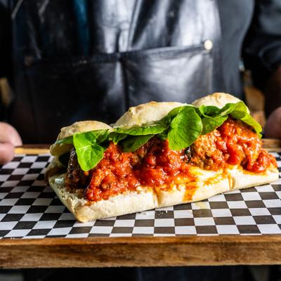 Person holding a tray with Meatball Sub sandwich topped with basil leafs.