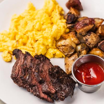 Steak and scrambled eggs, served with home fries and a small cup of ketchup.
