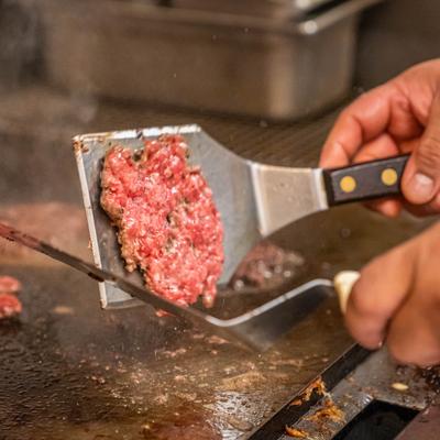 A person cooking a patty on a griddle with a spatula and tongs.