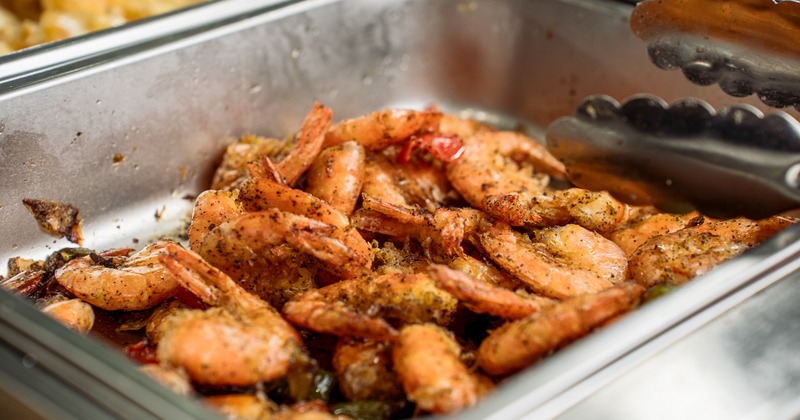 Seasoned peel-on shrimp fill a metal buffet pan under serving tongs