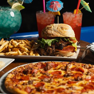 A table with a pepperoni pizza, a loaded burger with fries, and colorful cocktails with umbrellas.