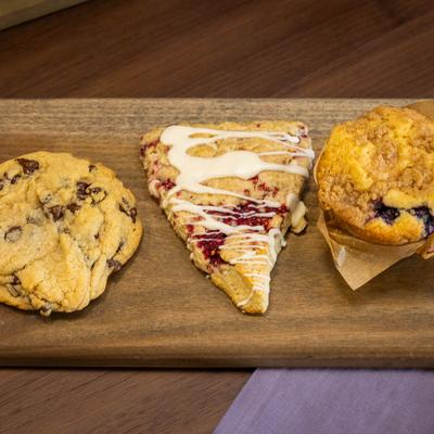 A wooden board holds a chocolate chip cookie, white chocolate raspberry scone, and blueberry muffin.