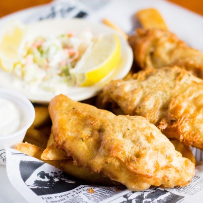 Fried fish pieces on top of chip style french fresh next to a side of coleslaw, lemon and tartar sauce