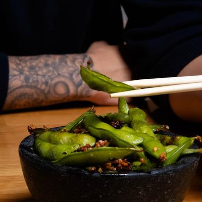 Chopsticks holding a seasoned edamame pod above a black bowl filled with more pods.