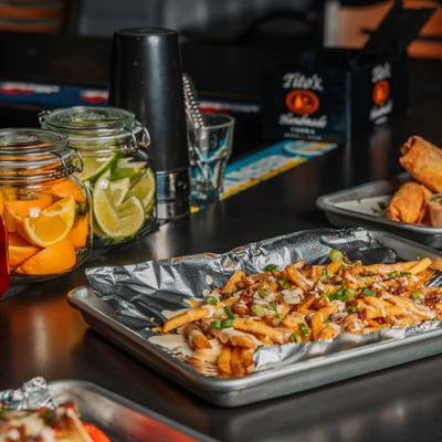 Loaded bacon fries on a bar counter surrounded by jars of citrus garnish and street rolls.