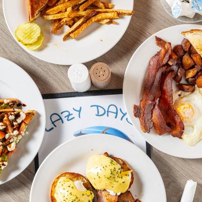 Multiple food plates sit on a wooden table with Lazy Days menu, overhead view.