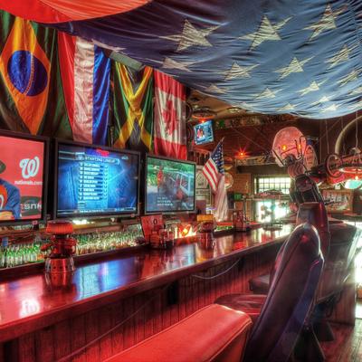 A lively bar with international flags, three TVs showing sports, a wooden counter, and bar stools