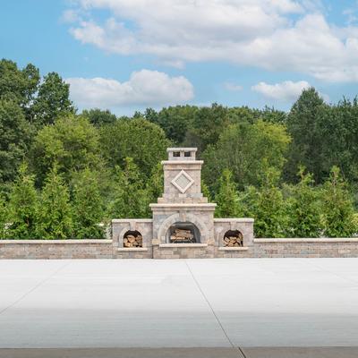 Outdoor stone patio with a stone fireplace and greenery behind it.