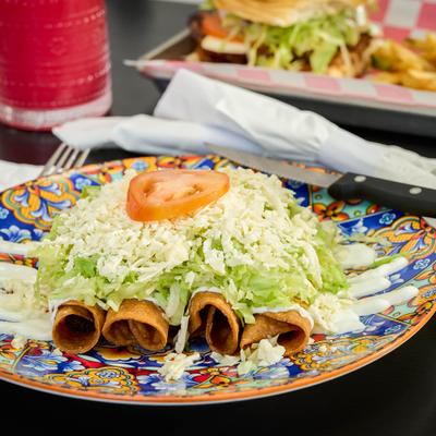 Four rolled tacos topped with shredded lettuce, white cheese, and tomato slice.