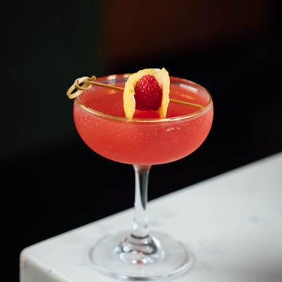 Red cocktail in a coupe glass, lemon peel and raspberry garnish