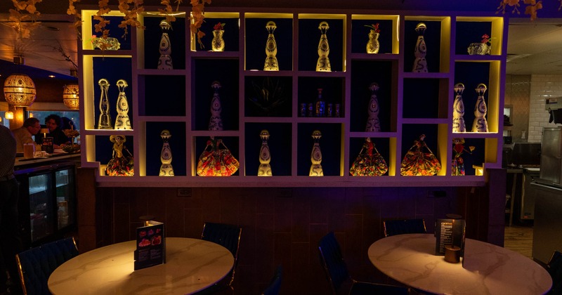 Dimly lit restaurant with shelves displaying dressed tequila bottles, tables and chairs