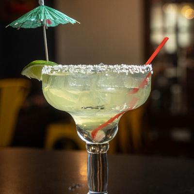 A glass of margarita with salted rim, lime, and a green umbrella, sits on a dark table.