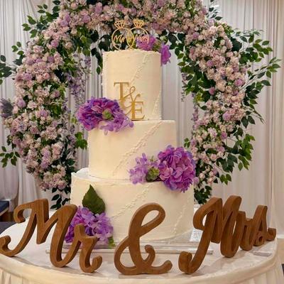 Tiered white wedding cake decorated with purple flowers, gold initials, and a wedding ring topper.