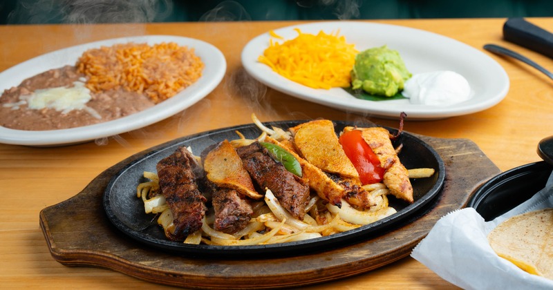 Combo fajitas served in a skillet on a wooden base and separate plates of sides