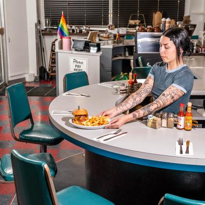 Interior, an employee  serving a burger and fries on a round counter with retro stools.