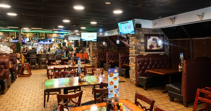 Interior of a warmly lit restaurant with wooden tables, leather booths, a stone bar area, and TVs showing sports