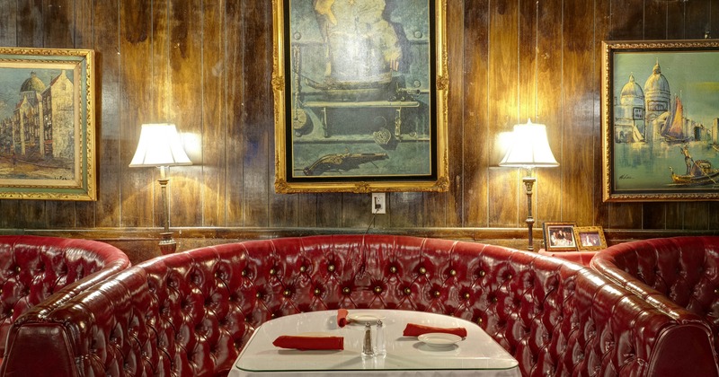 Red leather seating booth, table - white cloth and glass top;  lamps on a wooden wall with paintings