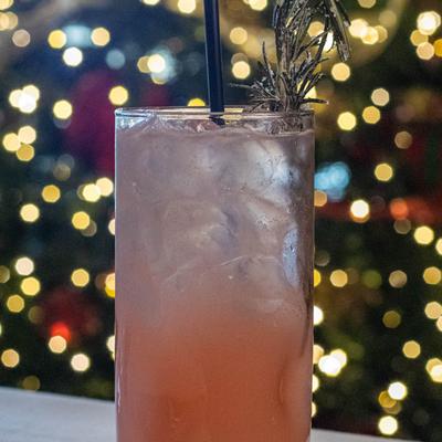 A tall glass of pink iced cocktail with a black straw and rosemary garnish.