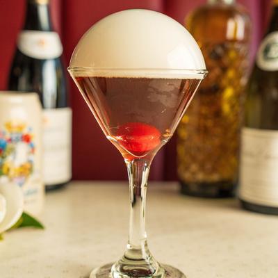 A red cocktail served in a martini glass, with foam dome on top.