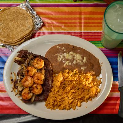 Steak topped with shrimp, served with rice, beans, tortillas, and iced drink.