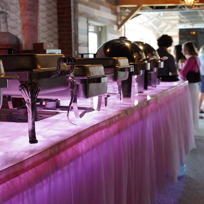 Chafing dishes sit on a glowing purple buffet table at an event.