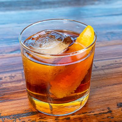 Old Fashioned cocktail served over large ice cube, garnished with orange peel.