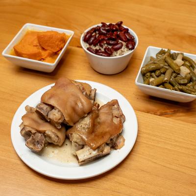 A plate of pigs feet served with bowls of sweet potatoes, red beans and rice, and green beans.