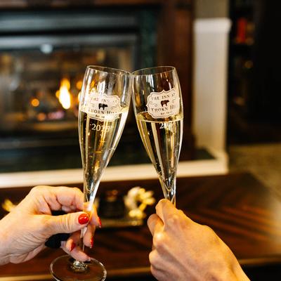 Two people clinking champagne flutes in front of a lit fireplace.