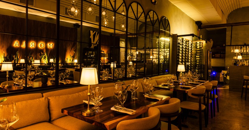 Warmly lit restaurant interior with tables, chairs, a mirror wall, and wine racks