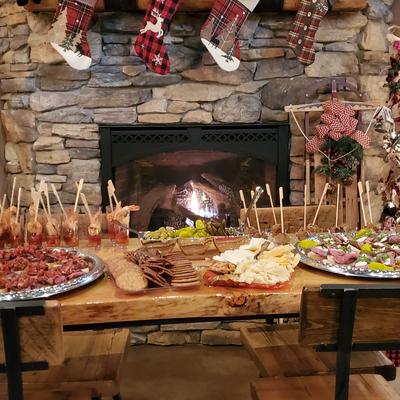 A wooden table holds appetizer platters and snacks in font of a lit stone fireplace.