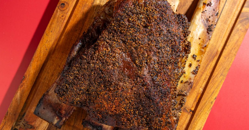 Close-up of a seasoned, grilled beef rib  on a wooden board against a red background