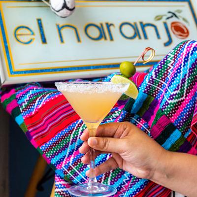 A hand holding a cocktail with lime and olive garnish in front of the El Naranjo restaurant sign.