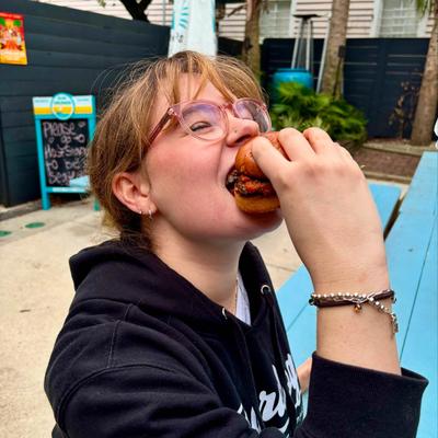 A person joyfully bites into a burger on a blue bench outdoors.