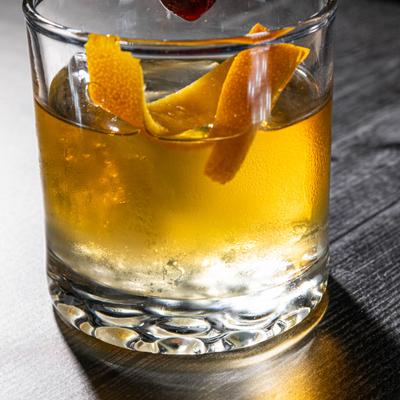 A chilled cocktail with an orange twist in a clear glass, topped with a cherry on a skewer.
