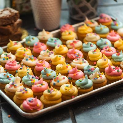 Colorful mini cupcakes with pink, yellow, and blue frosting topped with sprinkles fill a tray.