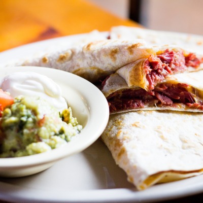 Quesadilla stuffed with corned beef on a plate served with a dish that contains guacamole, sour cream and pico de gallo