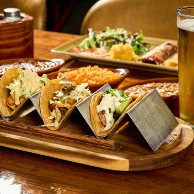 Portion of tacos served on the table with sides, enchillada platter and a glass of beer.