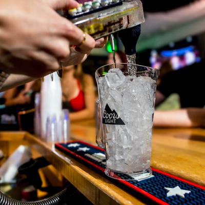A bartender pours a drink into a glass filled with ice at a bar, with patrons visible in the background.