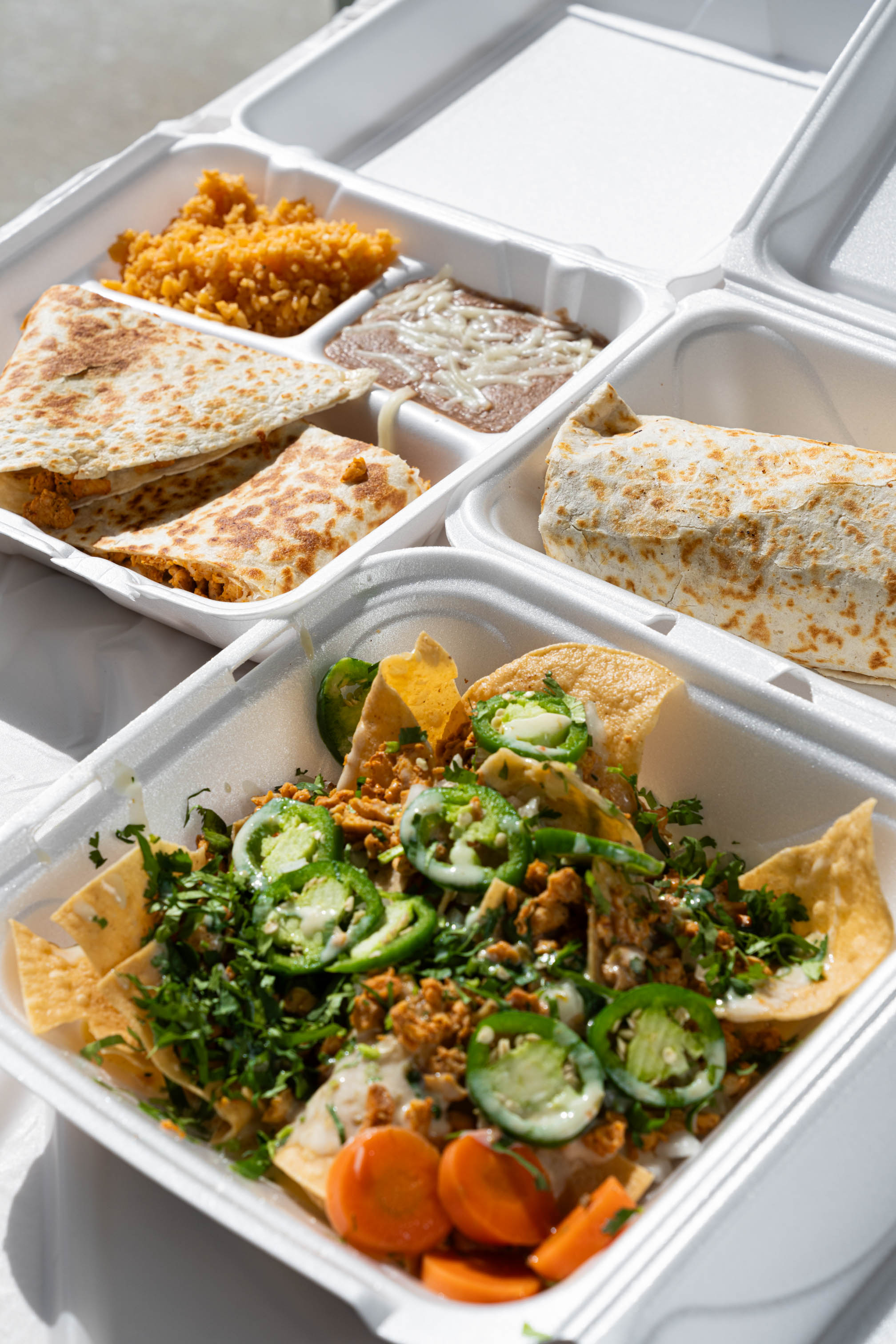 Loaded nachos topped with jalapenos, burrito, and slices quesadilla in takeout containers
