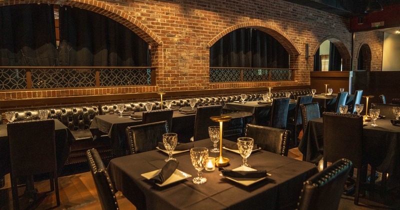 Interior of an elegant restaurant with black tablecloths, glassware on tables, and tufted black seating against a brick wall