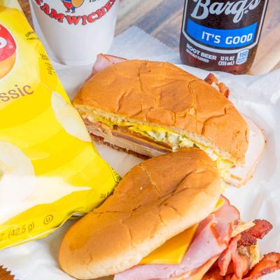 Two sandwiches on a table with a bag of chips and a root beer.
