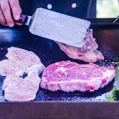 A hand pressing down on a steak with a spatula on a grill.
