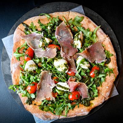 Pizza with burrata cheese, arugula, pesto, grape tomatoes, and prosciutto.