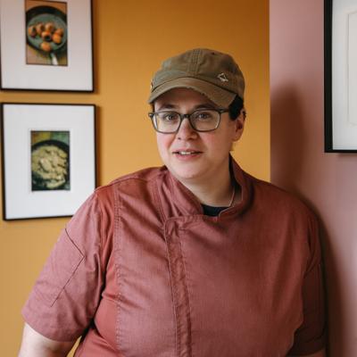 Chef in a pink jacket and cap standing by framed food photos.