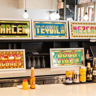 Colorful signs reading Tequila, Mezcal, and Good Vibes behind the counter with drinks.