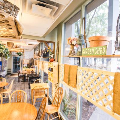Interior featuring wooden tables, chairs, and large windows with plants and shelves.