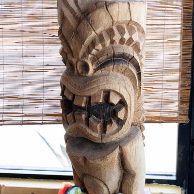 Interior, an ornate wooden tiki statue in front of a bamboo window blind.