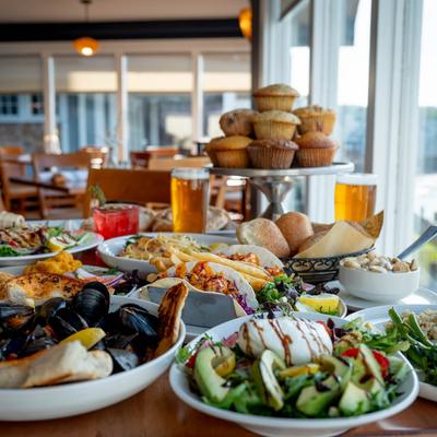 A lavish spread of assorted foods covers a wooden table in a bright restaurant.