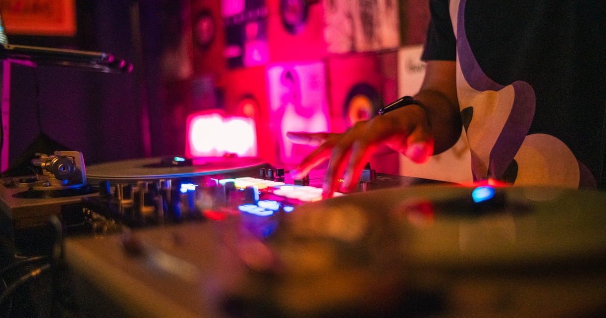 A DJ’s hand adjusts controls on a turntable in a dimly lit space with vibrant pink and blue neon lights