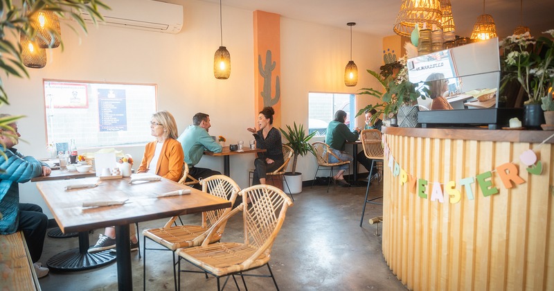Cafe interior with warm lighting, rattan chairs, and plants, Patrons are conversing at wooden tables